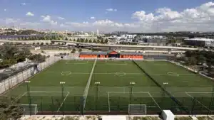 Yüksekten çekilmiş, yan yana üç suni çim halı saha; sahalar arasında fileler, ön tarafta aydınlatma direkleri ve kaleler, arkada turuncu tesis binası, köprülü yol ve uzakta lunapark rayları