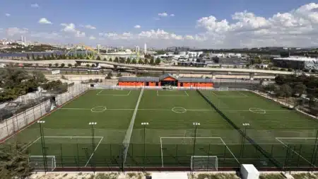Yüksekten çekilmiş, yan yana üç suni çim halı saha; sahalar arasında fileler, ön tarafta aydınlatma direkleri ve kaleler, arkada turuncu tesis binası, köprülü yol ve uzakta lunapark rayları