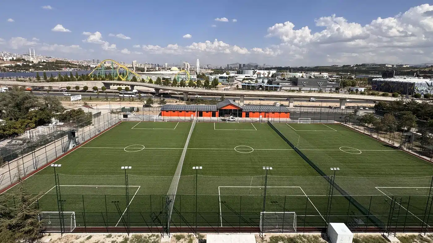 Yüksekten çekilmiş, yan yana üç suni çim halı saha; sahalar arasında fileler, ön tarafta aydınlatma direkleri ve kaleler, arkada turuncu tesis binası, köprülü yol ve uzakta lunapark rayları