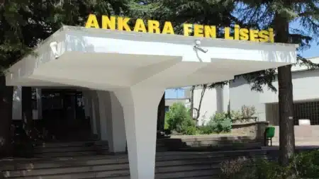 Ankara Fen Lisesi’nin beyaz betonarme giriş saçağı; üstünde sarı büyük harflerle “ANKARA FEN LİSESİ” yazısı, altında geniş basamaklar ve çevrede çam ağaçları