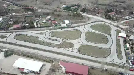 Havadan çekilmiş açık hava karting pisti: çimlik adacıkların çevresinde kıvrımlı, lastik bariyerli gri asfalt pistte dönen birkaç kart; önde yol ve kırmızı, beyaz çatılı yapılar, sağda park hâlindeki arabalar, arkada bahçeli tek katlı evler ve kurak tepeler