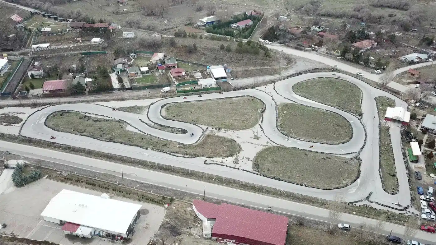 Havadan çekilmiş açık hava karting pisti: çimlik adacıkların çevresinde kıvrımlı, lastik bariyerli gri asfalt pistte dönen birkaç kart; önde yol ve kırmızı, beyaz çatılı yapılar, sağda park hâlindeki arabalar, arkada bahçeli tek katlı evler ve kurak tepeler