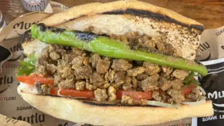 Yakından ciğer ekmek arası: közlenmiş ekmekte kızarmış ciğer parçaları, domates, soğan ve közlenmiş uzun yeşil biber; altta “Sokak Lezzetleri” baskılı kâğıt