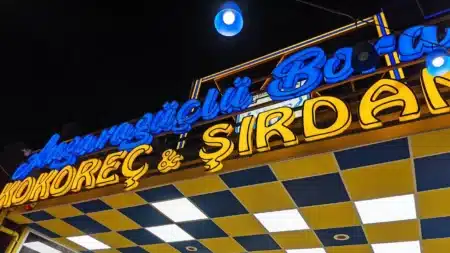 Gece aşağıdan çekilmiş ışıklı tabela: mavi el yazısıyla “Angaragüçlü Bora”, altında sarı büyük harflerle “KOKOREÇ & ŞIRDAN”; önünde mavi-sarı dama desenli, panel ışıklı saçak tavanı ve mavi sarkıt lambalar
