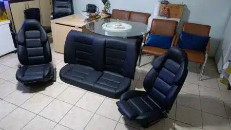 Oto döşeme atölyesinde yenilenmiş siyah deri araç koltukları: iki spor ön koltuk ve arka oturma minderi fayans zemin üzerinde, arkada masa ve kahverengi bekleme koltukları