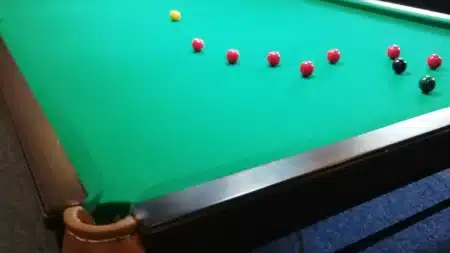Yeşil çuhalı snooker masası yakın çekim: sıra hâlinde kırmızı toplar, sarı, siyah ve yeşil top, ön planda kahverengi deri cep ve koyu ahşap masa kenarı