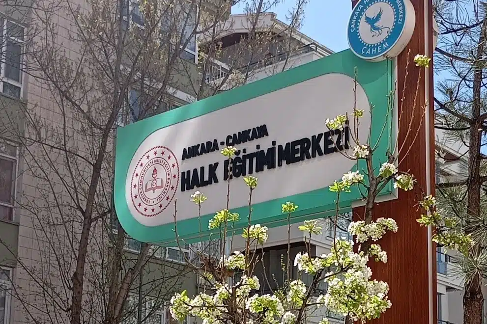 Ankara Çankaya Halk Eğitimi Merkezi’nin yeşil çerçeveli tabelası, MEB logosu ve CAHEM amblemi; önünde çiçek açmış bir ağaç