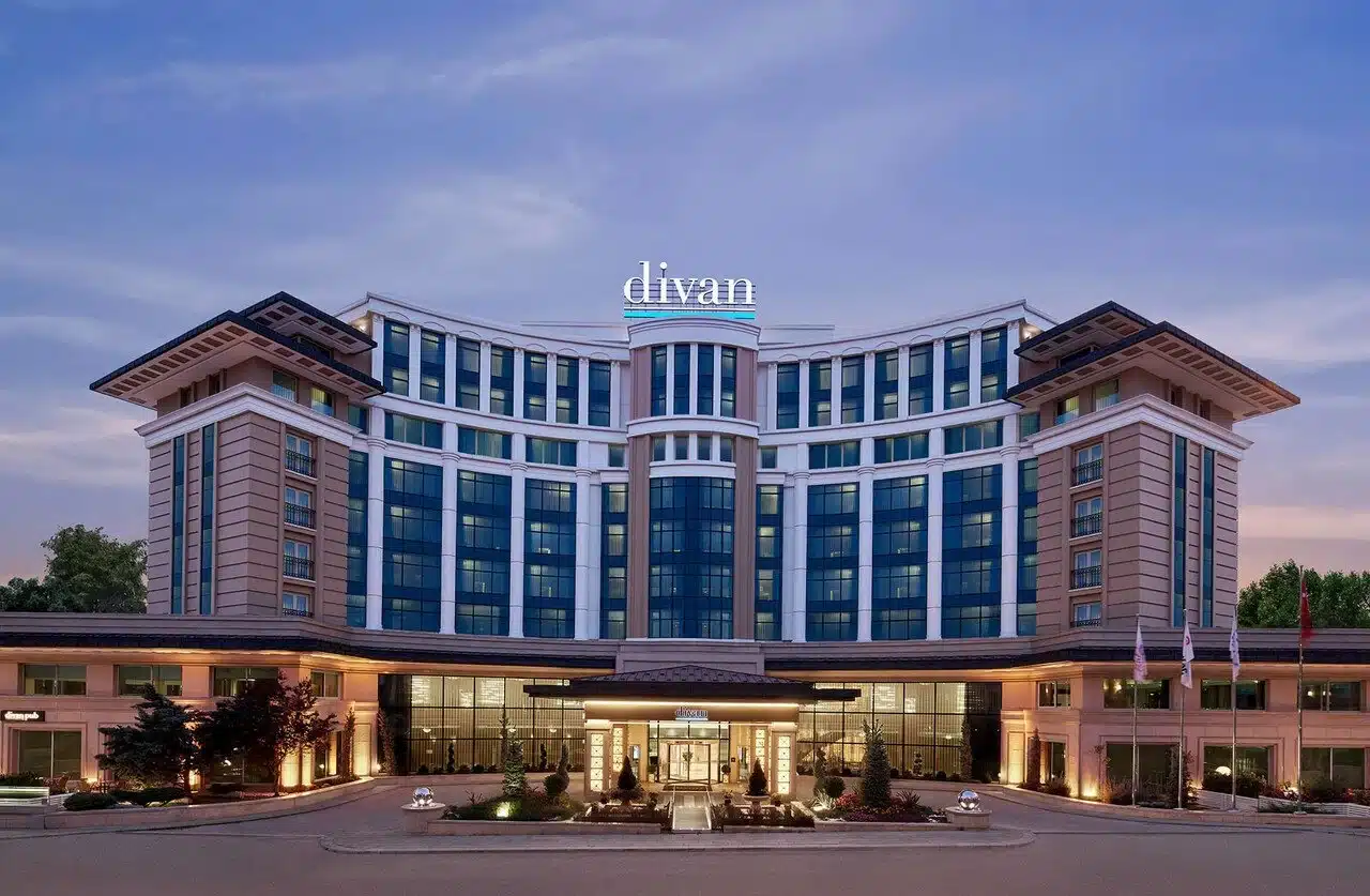 Divan Ankara otelinin alacakaranlıkta ışıklandırılmış cephesi: yarım daire planlı çok katlı bina, çatıda “divan” tabelası, önde aydınlatılmış giriş saçağı ve bayraklar