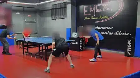 Kırmızı zeminli salonda mavi Stiga masa tenisi masasında karşılıklı oynayan oyuncular; arkadaki siyah duvarda “Emek Masa Tenisi Kulübü” logosu, sayı tabelası ve Stiga kenar panoları