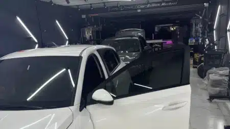 Emrah Demirel Oto Estetik Merkezi atölyesinde kapısı açık beyaz otomobil; ön kapı camında koyu cam filmi, arkada ikinci bir araç ve aydınlatmalı duvarlar