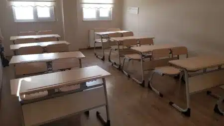Eryaman Tunafen Kurs’ta boş bir derslik: ahşap zeminde sıralanmış ikişerli bej sıralar ve sandalyeler, perdeli iki pencere, kalorifer ve duvarda askılık