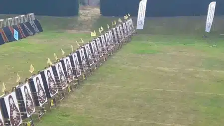 Çimenli açık hava okçuluk alanında çapraz uzanan uzun bir hedef hattı: numaralı ayaklar üzerinde armut biçimli geleneksel Türk okçuluğu hedefleri, hedeflere saplanmış oklar, her hedefin üstünde sarı bayrak; sağda Türkiye Geleneksel Türk Okçuluğu Federasyonu yazılı iki yelken bayrak