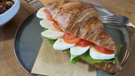 Gri seramik tabakta, kraft kâğıt peçete üzerinde kruvasan arasında mozzarella dilimleri, domates ve yeşil yapraklı sandviç; arkada çatallar, solda kuruyemişli bir kâse, ahşap masa
