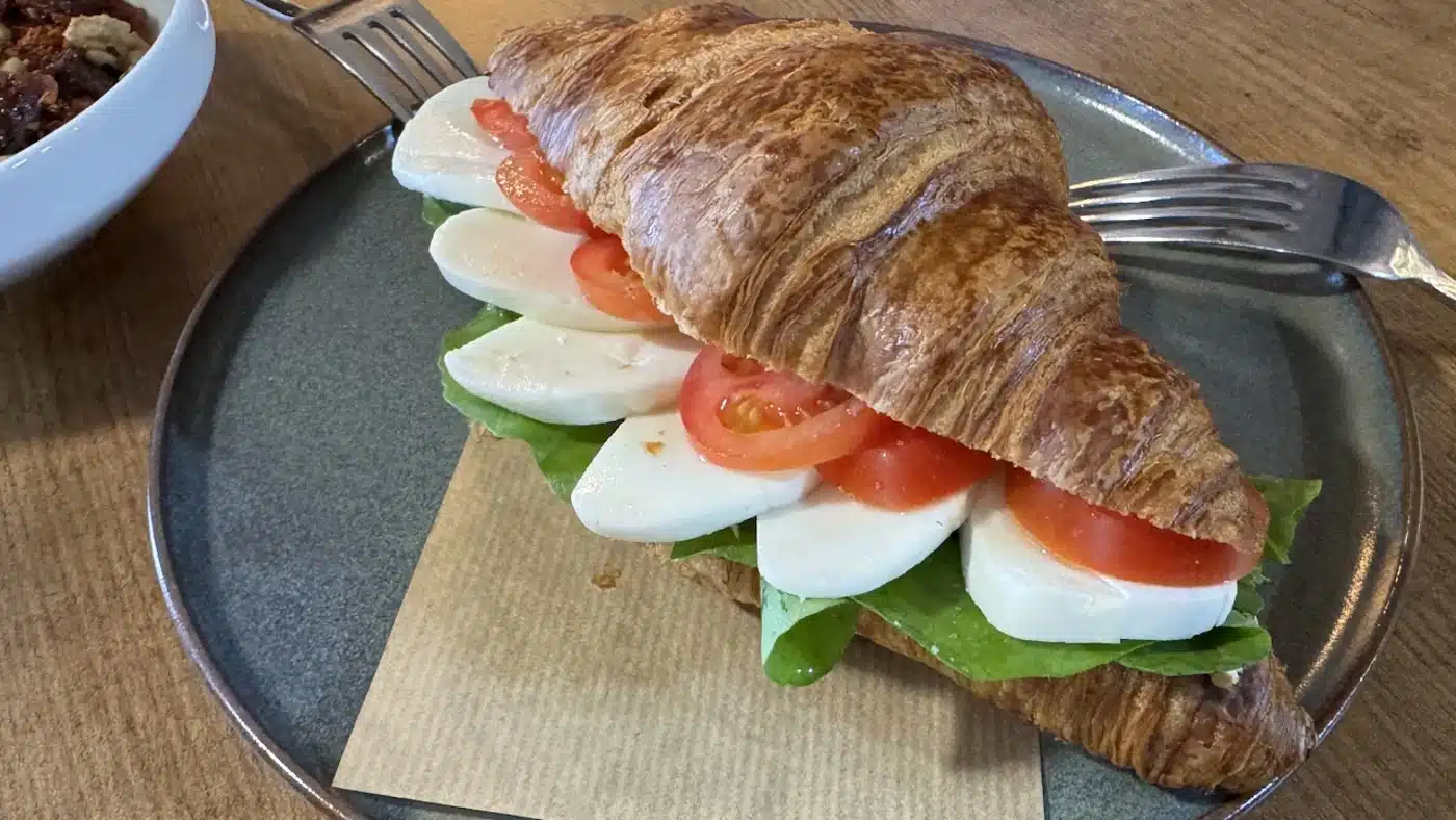 Gri seramik tabakta, kraft kâğıt peçete üzerinde kruvasan arasında mozzarella dilimleri, domates ve yeşil yapraklı sandviç; arkada çatallar, solda kuruyemişli bir kâse, ahşap masa