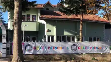 Güler Anaokulu’nun Mutlukent’teki binası: kiremit çatılı, açık yeşil boyalı iki katlı bina, önünde beyaz demir korkuluk boyunca renkli harflerle Güler Preschool yazılı afiş, ön planda çam ve sonbahar yapraklı ağaçlar