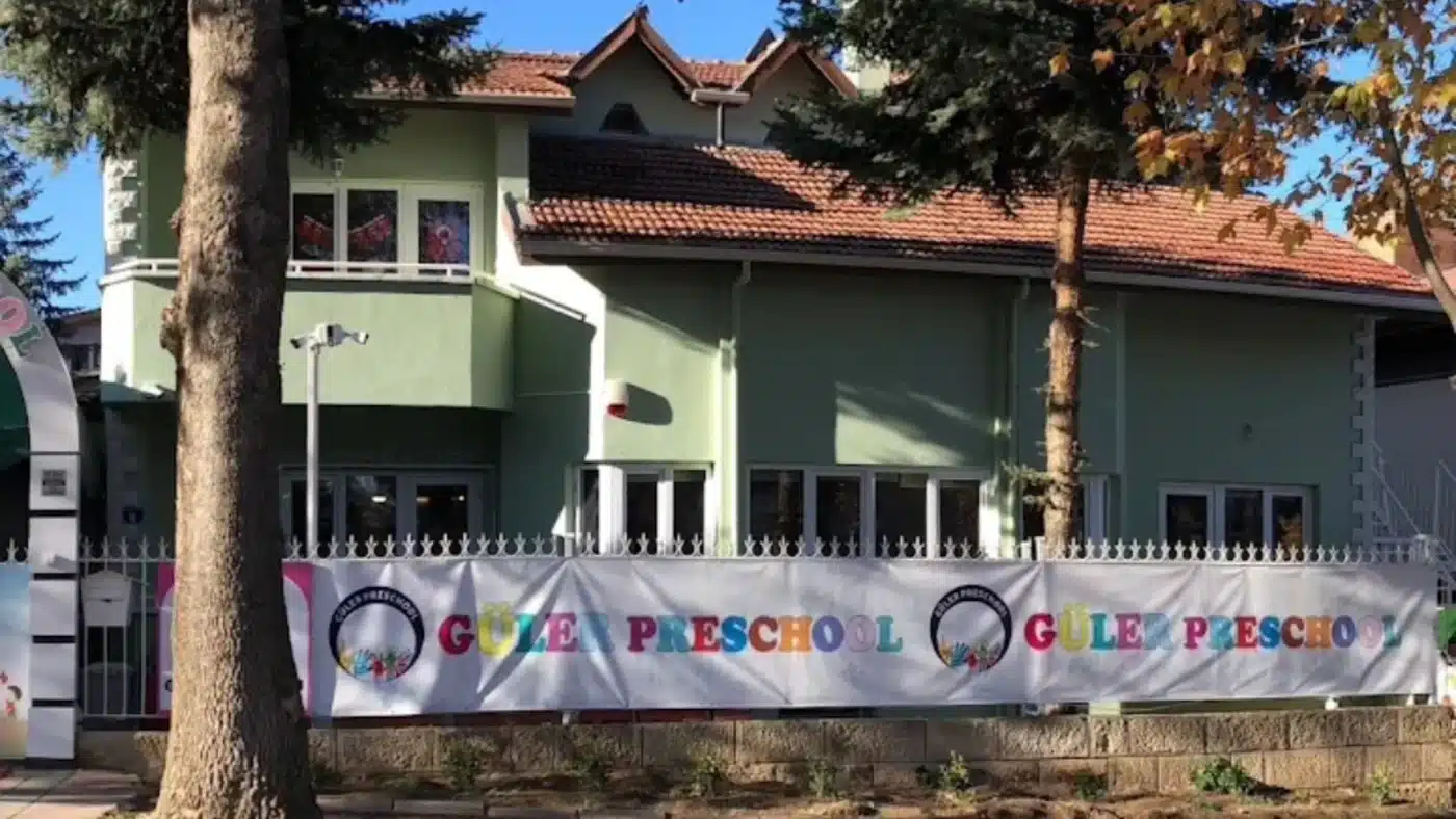 Güler Anaokulu’nun Mutlukent’teki binası: kiremit çatılı, açık yeşil boyalı iki katlı bina, önünde beyaz demir korkuluk boyunca renkli harflerle Güler Preschool yazılı afiş, ön planda çam ve sonbahar yapraklı ağaçlar