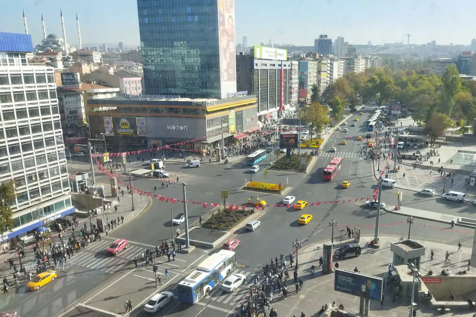 Kızılay AVM’nin üst katından Kızılay Meydanı: kavşak, metro girişi, sarı taksiler, EGO otobüsü, uzakta Kocatepe Camii minareleri ve sağda Güvenpark