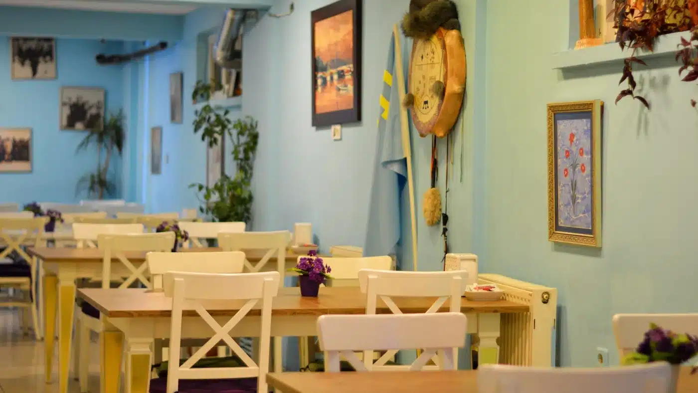Konakbay Çibörek Mantı Balgat’ın açık mavi duvarlı salonu: ahşap masalar, beyaz çapraz sırtlı sandalyeler, masalarda mor çiçekler; duvarda tablolar, açık mavi bir bayrak, kürk başlıklı davul biçimli süs ve çerçeveli ebru