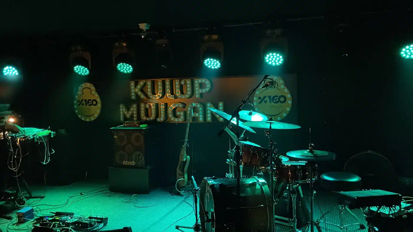 Kulüp Müjgan’ın konser sahnesi: arkada ampullü KULÜP MÜJGAN yazısı, yeşil sahne ışıkları altında davul seti, bas gitar, mikrofon ve bas amfisi; solda klavye ve efekt pedallarının başında bir müzisyen