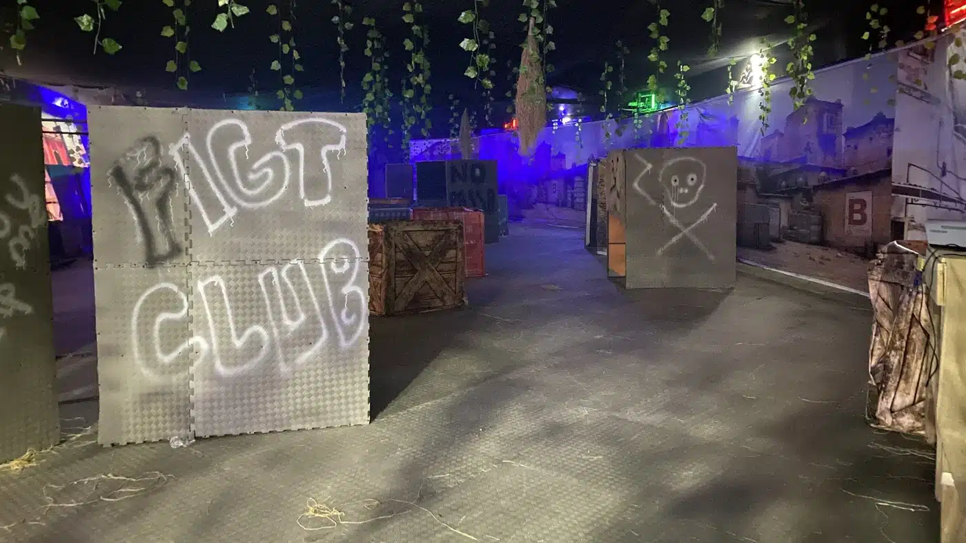 Loş, mor ve mavi ışıklı kapalı lazer tag arenası: solda “FIGT CLUB” yazılı grafitili siper paneli, ortada ahşap ve kırmızı sandıklar ile “NO PASSO” yazılı panel, sağda kurukafa grafitili siper, tavandan sarkan yapay sarmaşıklar ve duvarda şehir manzaralı dekor