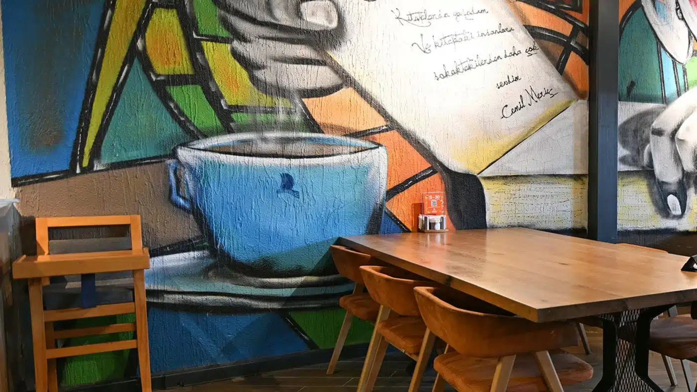 LİMAN Kitap Kahve’nin kafe salonu: ahşap uzun masa ve taba renkli sandalyeler, duvarda dumanı tüten mavi kahve fincanı, kitap sayfası tutan el resmi ve Cemil Meriç’in “Kitaplarda yaşadım ve kitaptaki insanları sokaktakilerden daha çok sevdim” sözü