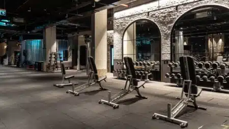 Ankara’nın En İyi Spor Salonları (Güncel Liste) MACFit Tunalı’nın serbest ağırlık alanı: kauçuk zeminde sıralı ayarlanabilir ağırlık sehpaları, sağda kemerli aynaların önünde dambıl rafları, solda halter ve ağırlık rafı
