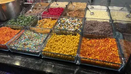 Müslüm Waffle’ın cam kaplarla dolu malzeme tezgâhı: turuncu, sarı ve rengârenk şeker toplar, damla çikolata, beyaz çikolata parçaları, kırmızı kokteyl vişne, portakal kabuğu ve yeşil kabak tatlısı şekerlemesi; solda waffle hamuru kazanı