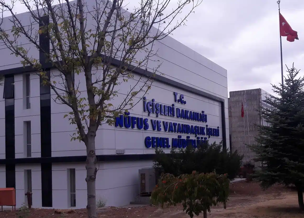 Nüfus ve Vatandaşlık İşleri Genel Müdürlüğü binası: beyaz cephede mavi harflerle “T.C. İçişleri Bakanlığı Nüfus ve Vatandaşlık İşleri Genel Müdürlüğü” yazısı, önde ağaçlar ve Türk bayrağı