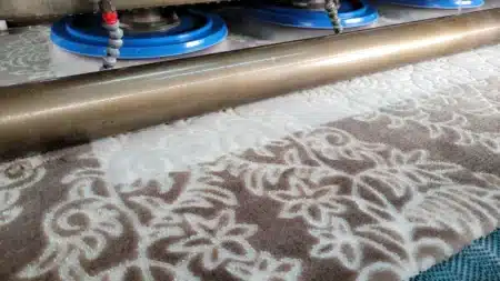 Halı yıkama makinesinde yıkanan krem zemin üzerine kahverengi kabartma desenli halı; üstte mavi döner fırça başlıkları ve su püskürten borular, önde pirinç renkli silindir