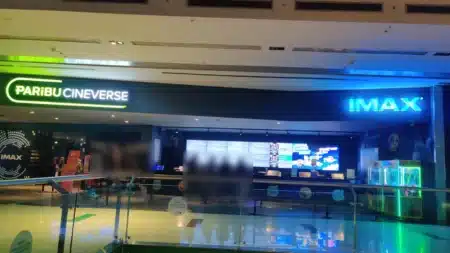 AVM koridorundan sinema girişi: solda yeşil ışıklı PARİBU CINEVERSE, sağda mavi ışıklı IMAX tabelası; ortada büyük dijital ekranlı gişe, sağda oyuncak makineleri, önde cam korkuluk