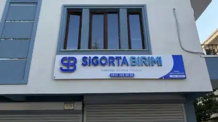 Mavi gri pencere çerçeveli açık renkli binanın cephesinde beyaz-mavi “Sigorta Birimi – Geleceğe Güvenle Yürüyün” tabelası, altında telefon numarası ve “Genel Merkez” yazısı; tabelanın üstünde ahşap çerçeveli pencere, altta kapalı kepenkler