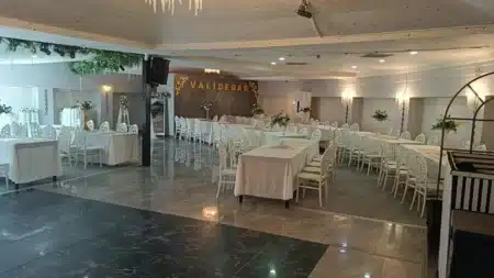 Validebağ’ın kapalı salonu: arka duvarda altın harflerle “VALİDEBAĞ” yazısı, beyaz örtülü uzun masalar, beyaz oymalı sandalyeler, masalarda çiçek aranjmanları, tavanda kristal avize ve önde mermer görünümlü boş dans alanı