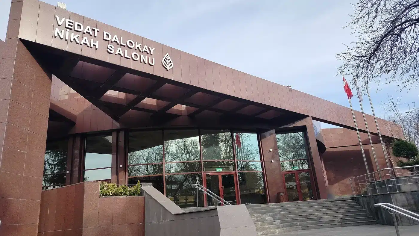 Kırmızı granit kaplı Vedat Dalokay Nikah Salonu binasının girişi: cephede “VEDAT DALOKAY NİKAH SALONU” yazısı ve Çankaya Belediyesi yaprak amblemi, camlı giriş kapıları, merdivenler ve bayrak direkleri